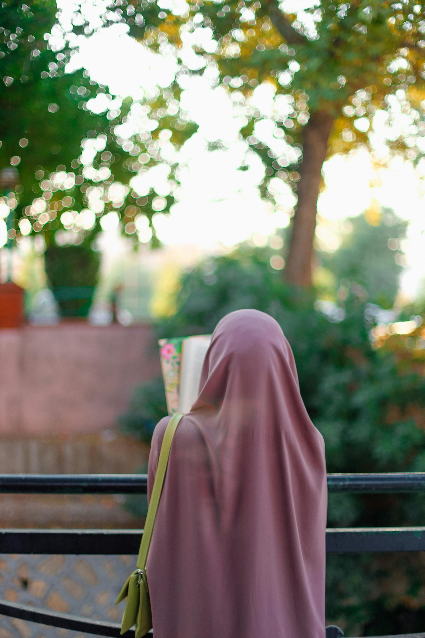 A woman in a hijab stands with her back to the camera, looking over a metal railing with a blurred outdoor background.