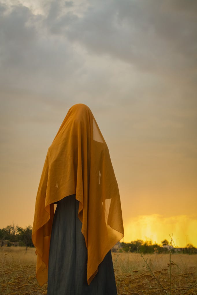 A woman in a yellow headscarf stands in a field at sunset, capturing a serene rural atmosphere.