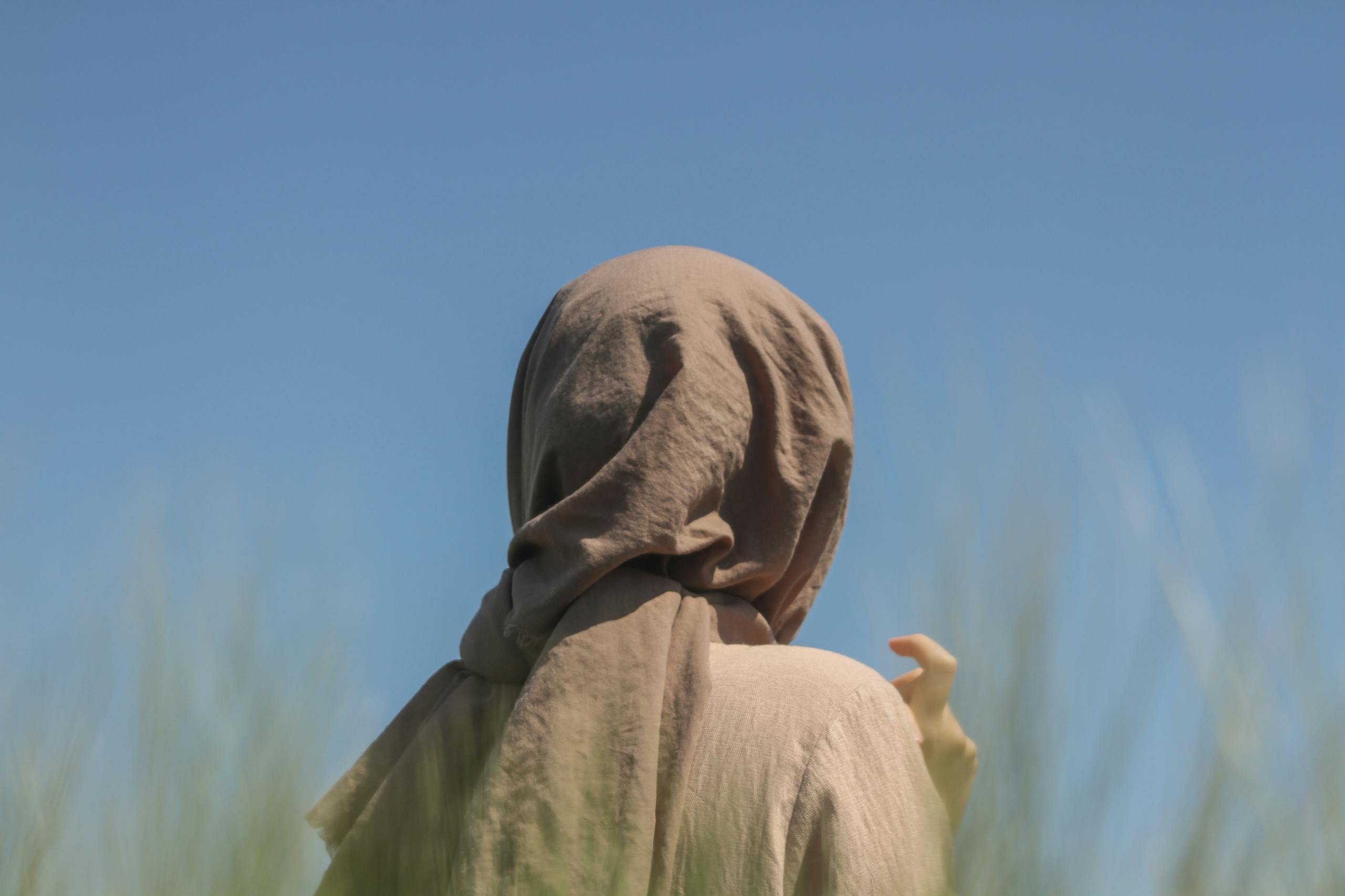 Person wearing a headscarf with a blue sky backdrop, captured from behind in an outdoor setting.