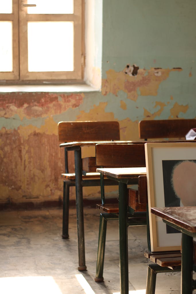 Sunlight spills onto vintage desks in an abandoned classroom, showcasing peeling walls and nostalgia.