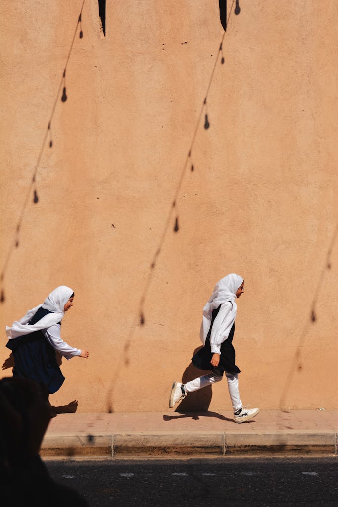Two young girls run energetically past a sun-dappled wall in Oman, capturing a moment of joyful childhood.